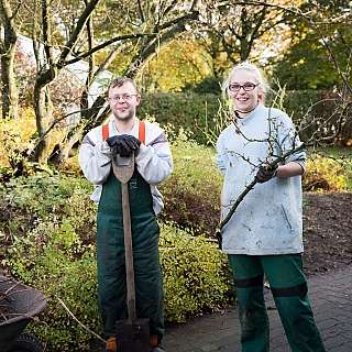 Zwei Gärtner halten Werkzeuge und Äste in einem Garten.